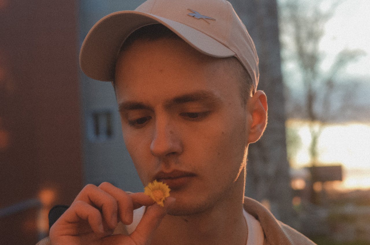 A man in a cap thoughtfully looks at a small yellow flower, capturing a moment of contemplation outdoors.
